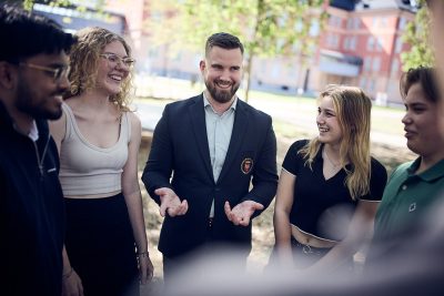 En lärare interagerar med studenter utomhus, alla ler och verkar engagerade.