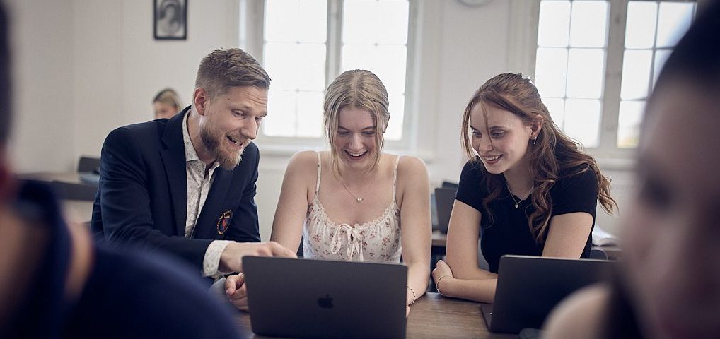 En lärare hjälper två studenter med datorarbete, alla skrattar.