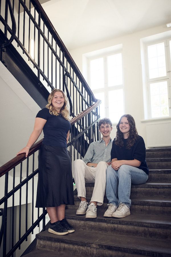 Tre elever hänger i trappan på skolan och ler in i kameran.