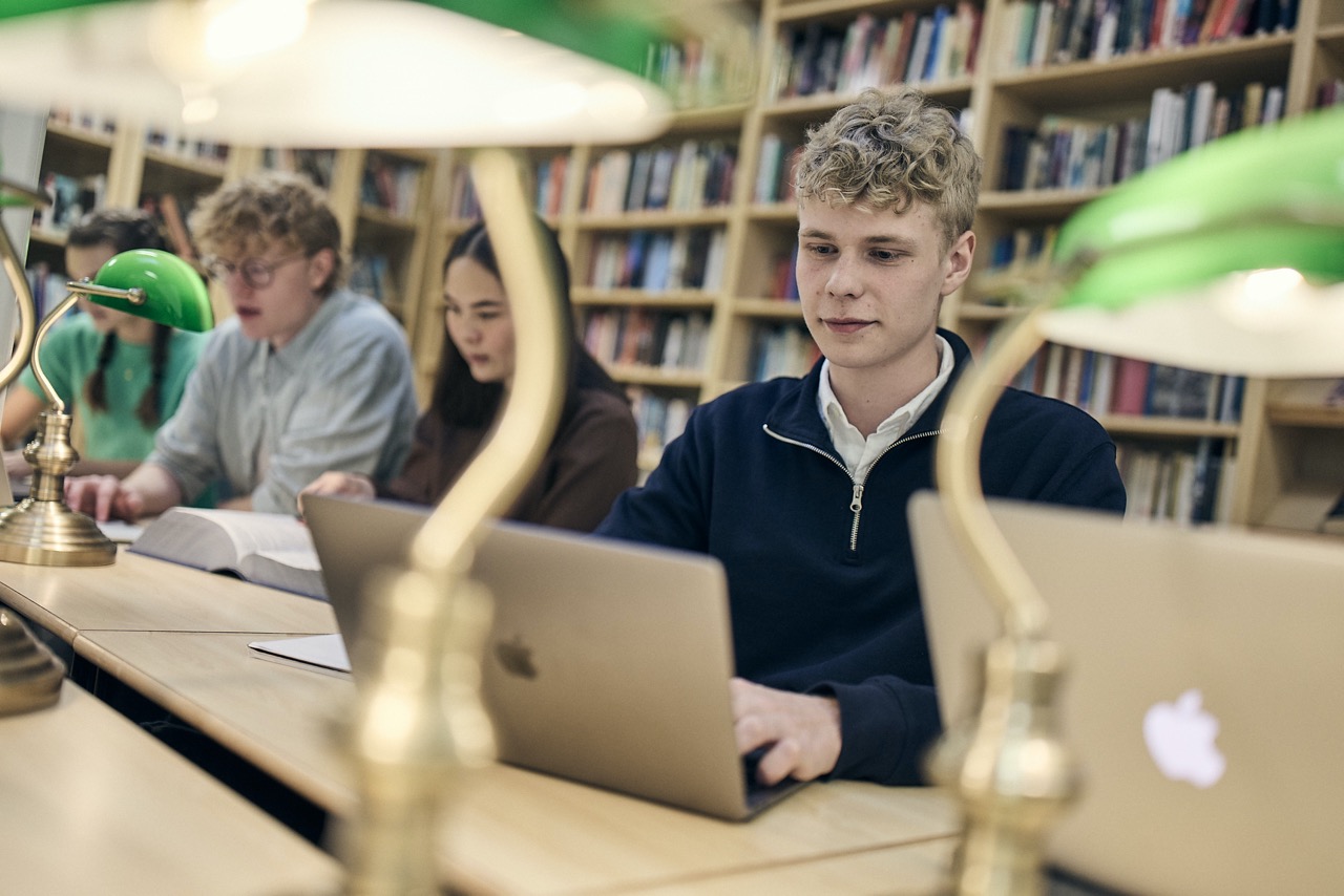 Elever sitter i skolbiblioteket och studerar med datorer och böcker.