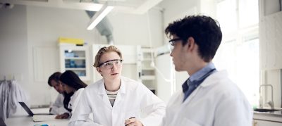 Två elever i labbrockar diskuterar och arbetar i ett kemilaboratorium.