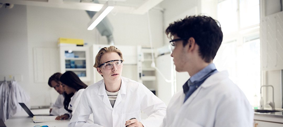 Två elever i labbrockar diskuterar och arbetar i ett kemilaboratorium.