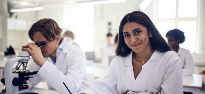 Elever i labbrockar arbetar i ett laboratorium, en elev använder mikroskop.